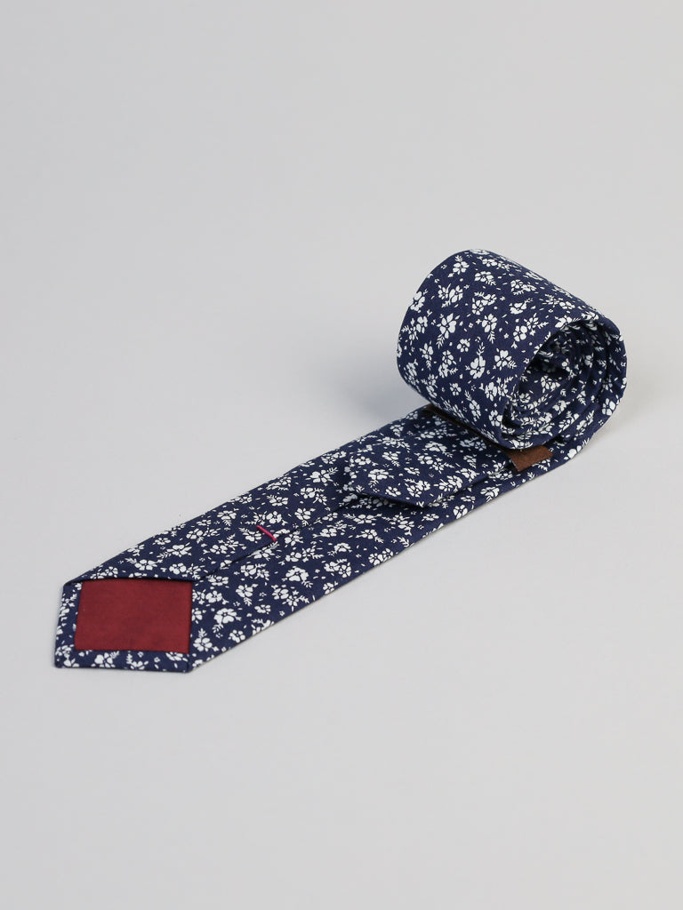 The Ditsy Cotton Tie by ID Menswear is a handmade navy blue cotton tie with a white floral pattern and a solid burgundy tip, neatly rolled at one end and displayed on a light gray background.