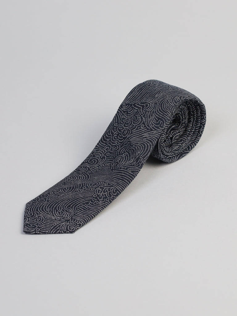 The Japanese Printed Nami Tie by ID Menswear features a navy blue base with a detailed white wave pattern. Handmade in Brooklyn, its rolled and shown on a light grey background—ideal for business formal occasions.