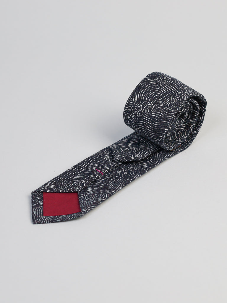 The Japanese Printed Nami Tie by ID Menswear, featuring a dark gray pattern and a red tip detail, is handmade in Brooklyn and displayed rolled on a light gray background.