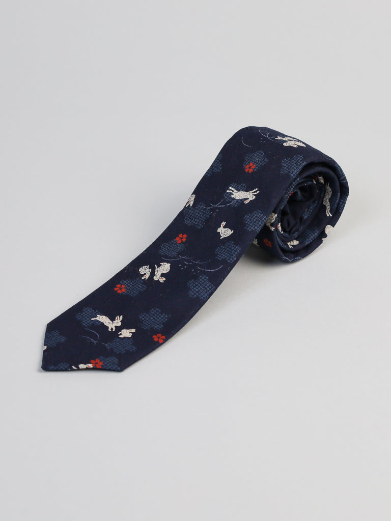 The Japanese Printed Bunny Tie by ID Menswear features white rabbits and red flowers on navy blue cotton. Handmade in Brooklyn, it’s shown rolled up on a light gray surface.