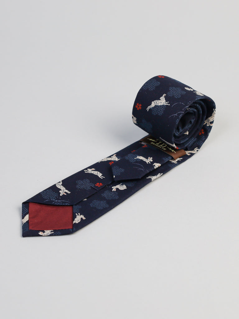 The Japanese Printed Bunny Tie by ID Menswear is navy blue with white cranes, red flowers, and clouds. Handmade in Brooklyn from 100% cotton, its shown on a light background, partially rolled to reveal a maroon back tip lining.
