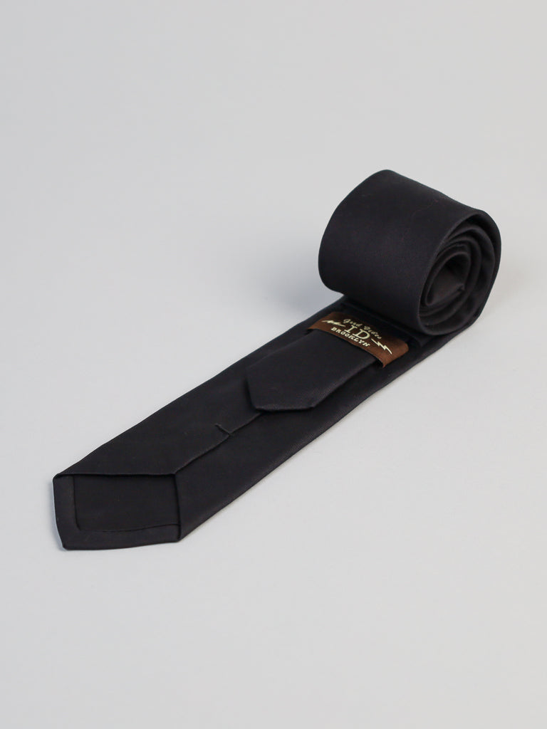 The New Black Cotton Tie by ID Menswear, handmade in Brooklyn, is neatly rolled and displayed on a light gray surface with a brown label featuring gold text on the back of its wide end.