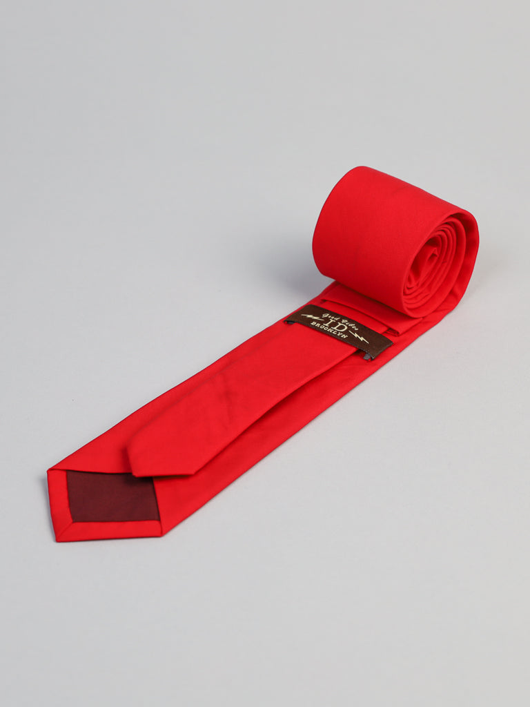 The Rouge Cotton Tie by ID Menswear, handmade in Brooklyn from bright red cotton, is shown rolled at one end and laid flat on a light gray surface, with its brown label visible inside the wider tip.