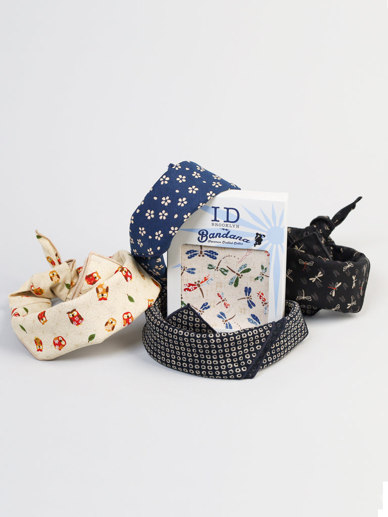 A set of four 100% cotton bandanas with assorted colors and prints is shown, including two cream with mushrooms, one blue with white flowers, and one black adorned with dragonflies. A card labeled Bandana is centrally placed. Product: Multi Colored Dragonfly Bandana by ID Menswear.