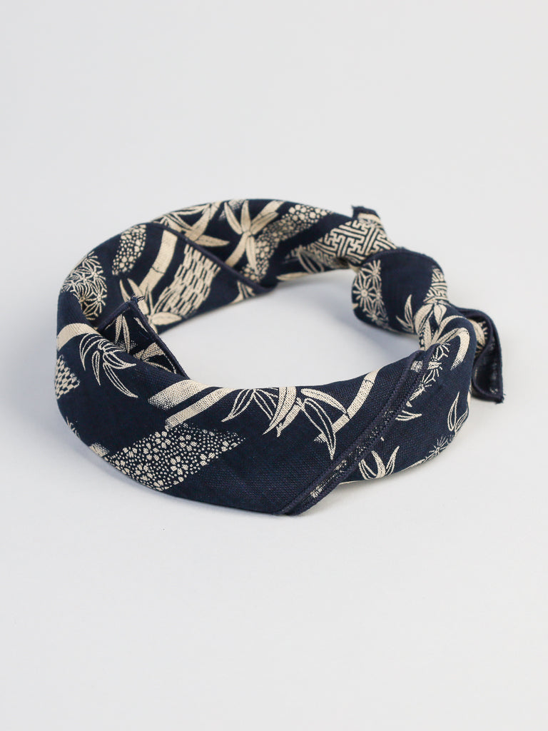 The Japanese Bamboo Bandana by ID Menswear, in navy blue with intricate beige patterns featuring geometric shapes and leaves, is crafted from 100% cotton. It elegantly twists into a loop against a white background.