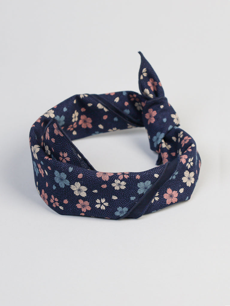 The Cherry Blossom Bandana by ID Menswear is a dark blue fabric headband with a knotted design. It features a floral pattern with pink and white cherry blossoms and is displayed on a light gray background, highlighting its elegance and charm.