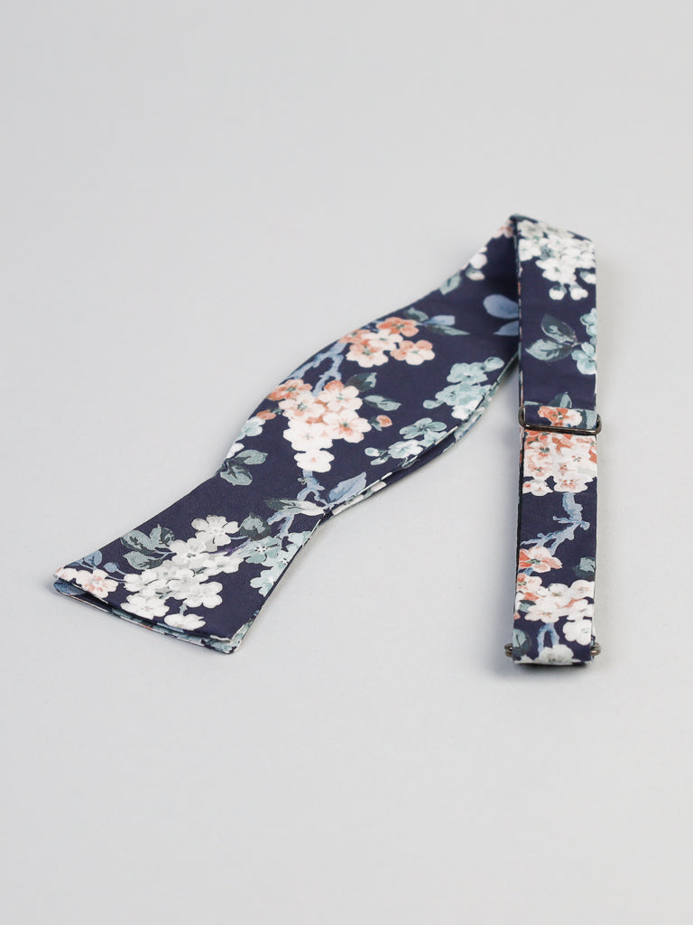 The ID Liberty of London Tana Lawn™ Josey Blue Cotton Bowtie by ID Menswear, handmade in Brooklyn, features a floral pattern with white and peach flowers. Untied on a light gray surface, it highlights its elegant design and texture.