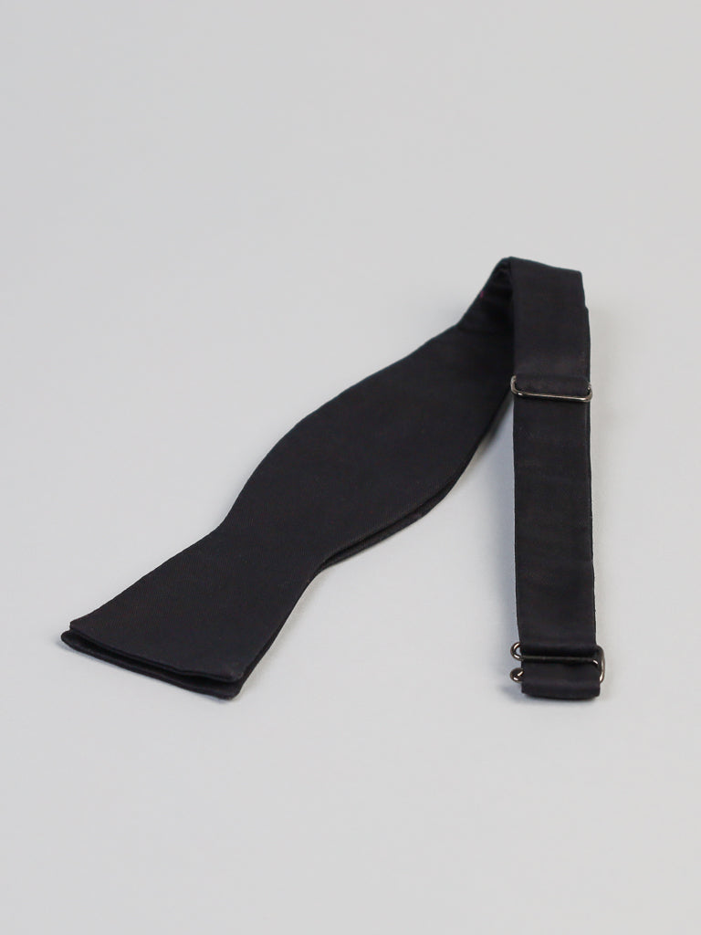 The New Black Cotton Bow Tie by ID Menswear is handmade in Brooklyn from smooth cotton, featuring an adjustable strap with a metal slider. It’s shown flat on a light gray surface, highlighting its classic design with wide and narrow ends.