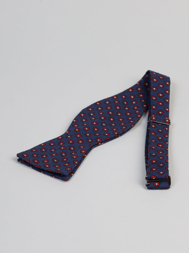 The Flower Dot Cotton Bow Tie by ID Menswear, handmade in Brooklyn, features a blue cotton fabric with a red and white circular pattern and is displayed flat on a light gray surface.