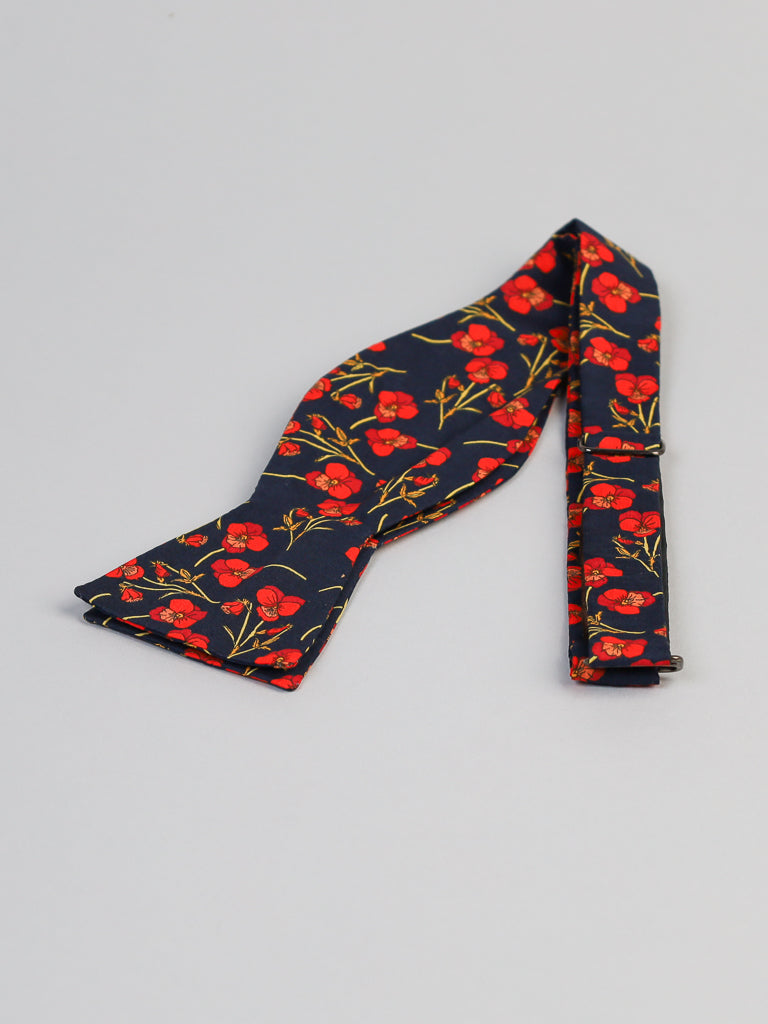 The ID Liberty of London Tana Lawn™ Poppy Red Cotton Bowtie by ID Menswear, featuring navy blue with vibrant red flowers and green stems, is displayed unfolded on a light gray background.