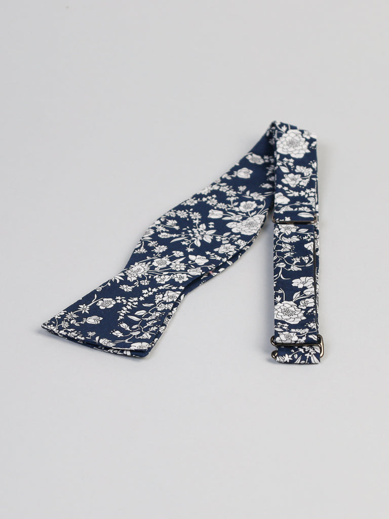 A bow tie from ID Menswear, crafted from Liberty of London Tana Lawn™ cotton, features a black background with a white floral pattern and is elegantly displayed on a light gray surface.