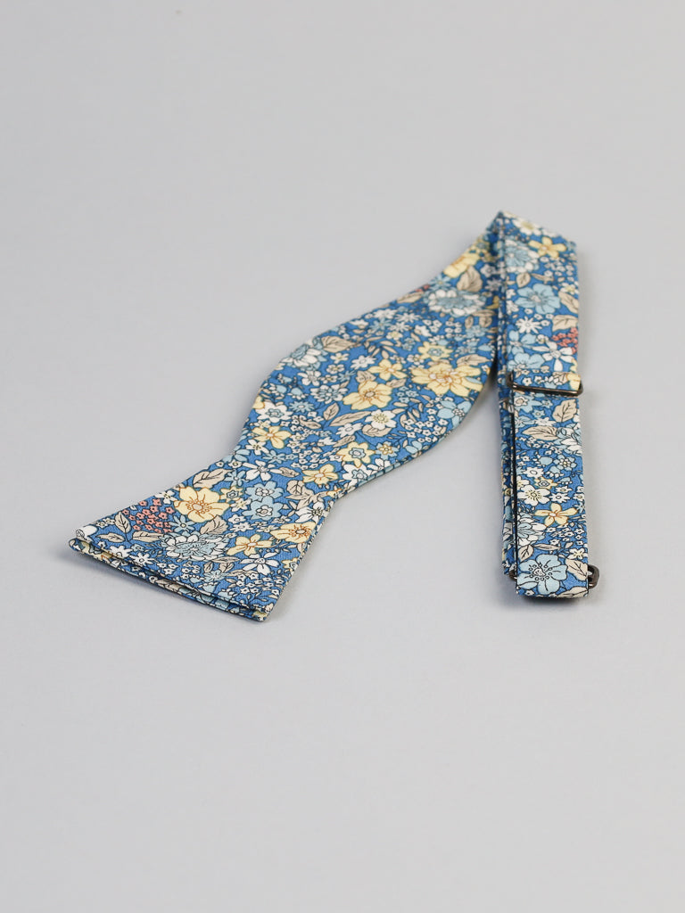 The Poplin Cotton Bow Tie by ID Menswear is a handmade, self-tie blue bow tie featuring yellow, white, and light blue florals in 100% cotton, shown flat on a light gray background.
