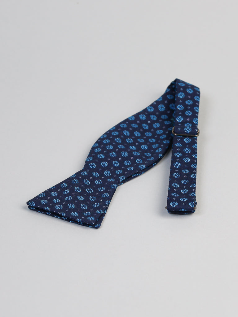 The Play Day Cotton Bow Tie by ID Menswear is handmade in Brooklyn from 100% cotton, showcasing a blue self-tie design with a small light-blue geometric floral pattern on a light gray background.
