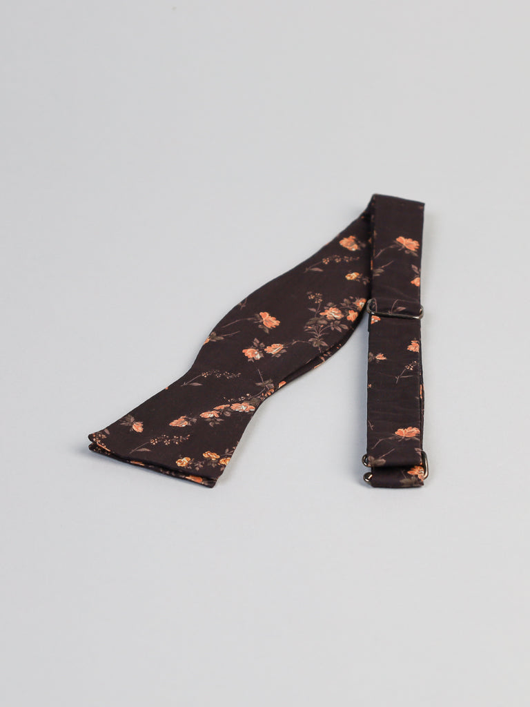The ID Liberty of London Tana Lawn™ Elizabeth Cotton Bowtie by ID Menswear, handmade in Brooklyn, is dark brown with a pattern of small orange flowers. It features an adjustable strap and is laid flat on a light gray background.