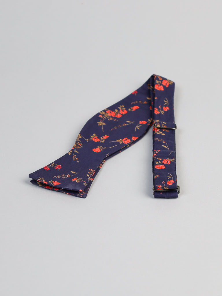 The ID Liberty of London Tana Lawn™ Elizabeth Cotton Bowtie from ID Menswear, featuring a dark blue fabric with red and orange floral patterns, rests on a light gray surface. Handmade in Brooklyn, this adjustable bow tie is presented unfolded.