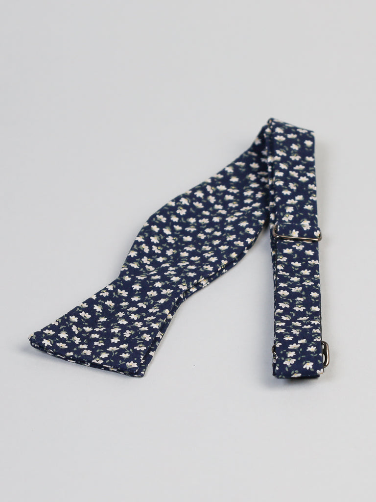 The Japanese Hana NW Printed Bow Tie by ID Menswear, handmade in Brooklyn from navy blue cotton with a white Japanese floral print, is shown laid flat on a light grey surface.