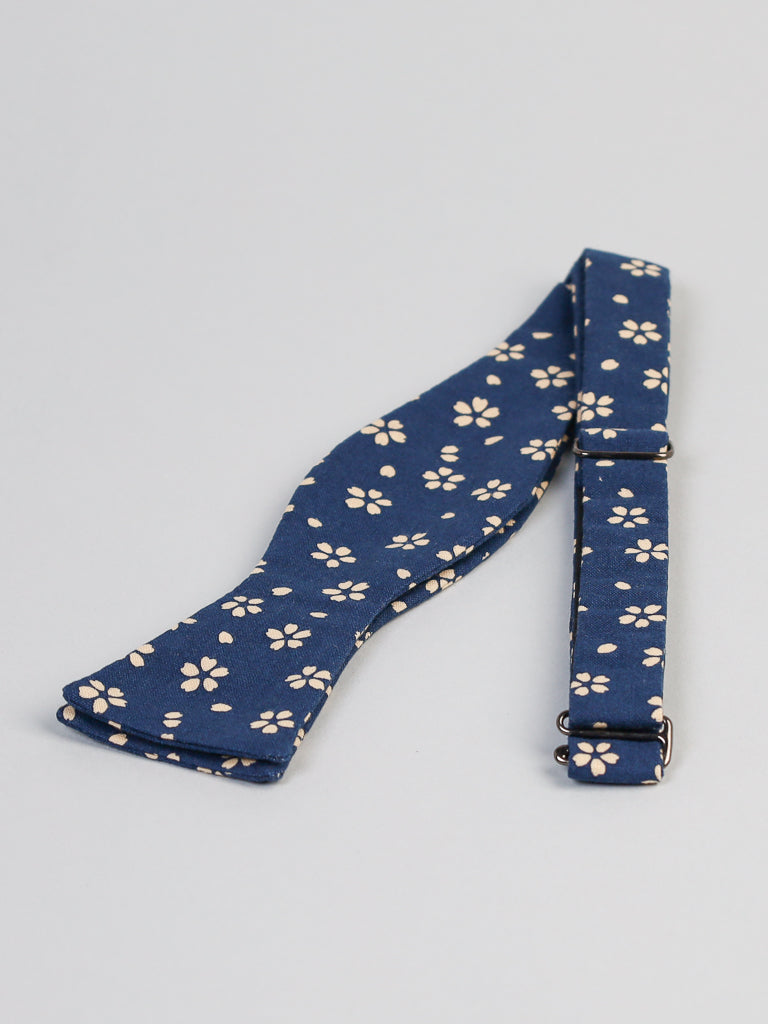 The Japanese Sakura Printed Bow Tie by ID Menswear is handmade in Brooklyn from 100% cotton Japanese fabric, featuring a blue floral print and an adjustable strap with metal clasps. Shown laid flat on a light gray surface.