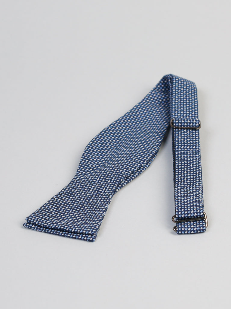 The ID Menswear Japanese Ya Cotton Printed Bow Tie, featuring a blue and white checkered pattern, is elegantly displayed on a light gray surface. Handmade in Brooklyn with a textured cotton fabric and adjustable metal clasp, it adds artisanal charm to your attire.