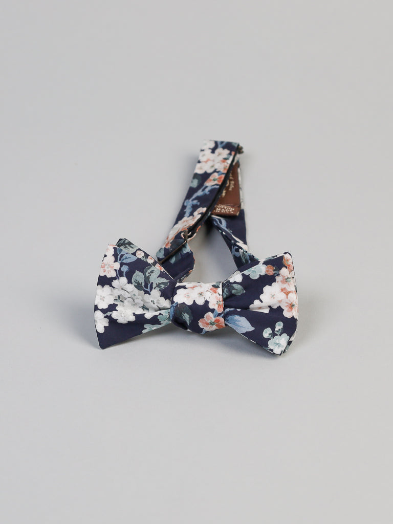 The ID Liberty of London Tana Lawn™ Josey Blue Cotton Bowtie by ID Menswear is handmade in Brooklyn. It showcases a floral pattern with navy, white, pink, and light blue on a gray background. The strap fits neatly behind the bow.