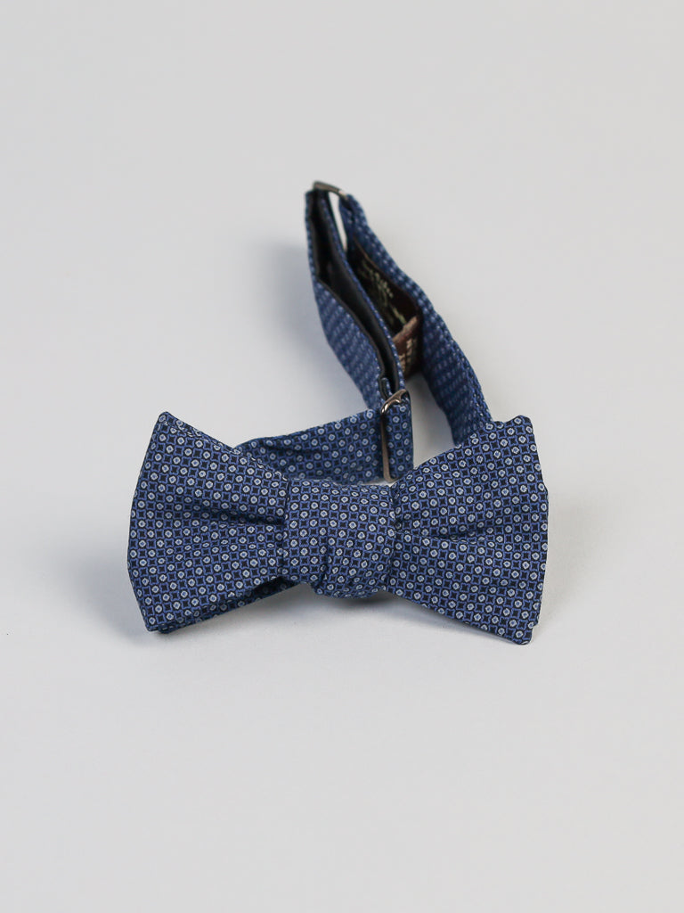 The Japanese Fleur Printed Bow Tie by ID Menswear features a blue pattern with small white dots, is handmade in Brooklyn from Japanese-printed cotton, pre-tied, includes an adjustable strap, and is shown on a plain light gray background.