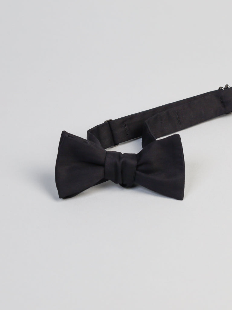 The New Black Cotton Bow Tie by ID Menswear, featuring an adjustable strap and handmade in Brooklyn, is displayed flat on a light gray surface.