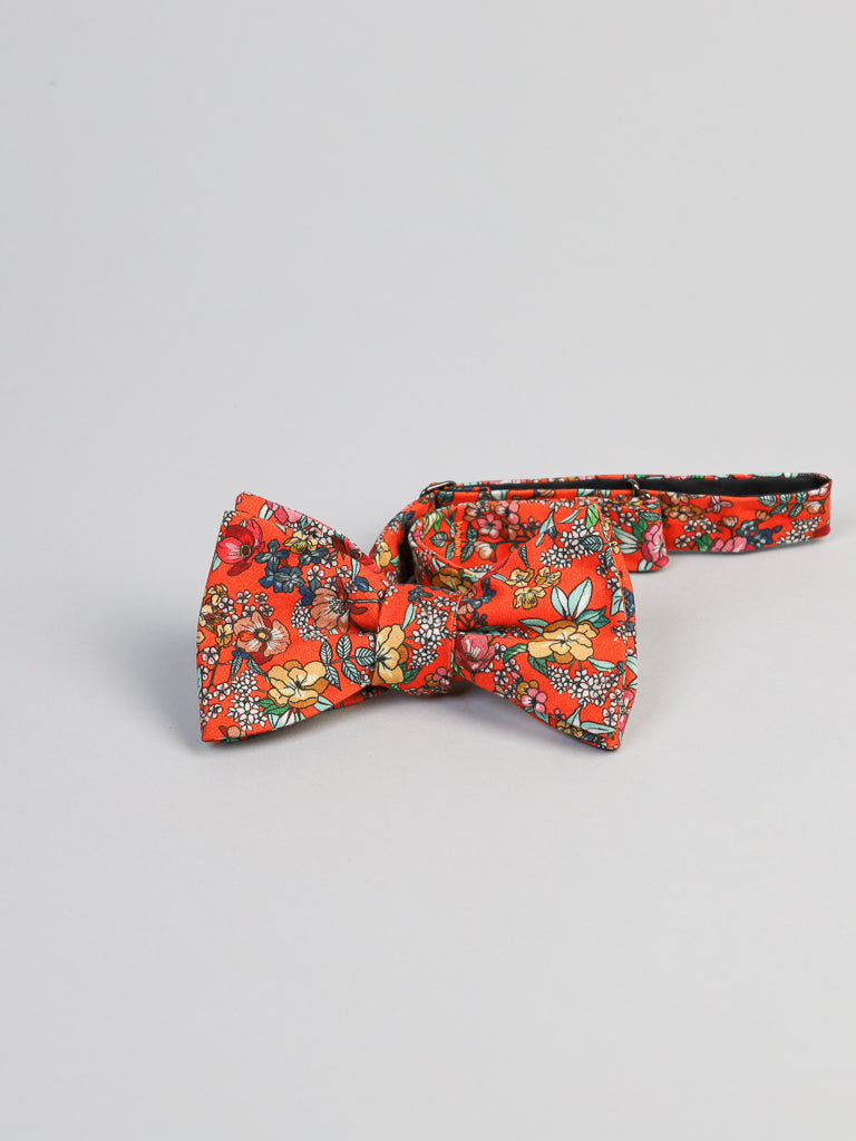 The Bouquet Cotton Bow Tie by ID Menswear features a red cotton fabric with yellow, white, and green floral patterns. Handmade in Brooklyn, it’s an ideal semi-formal accessory, shown here on a plain light background.