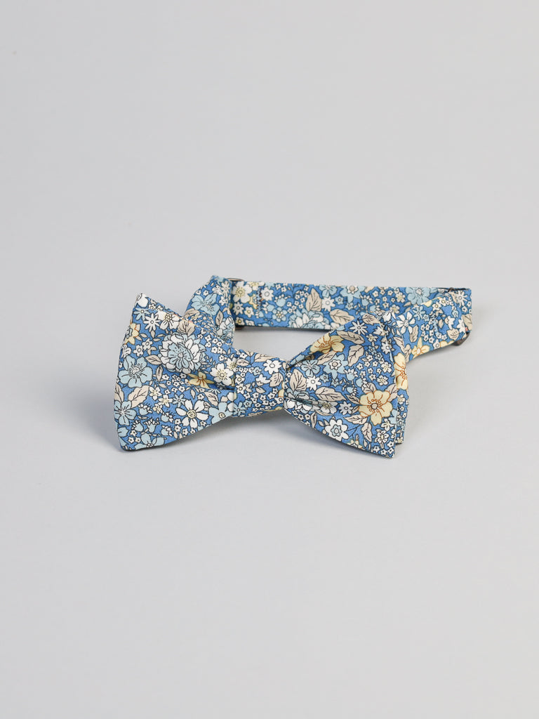 The Poplin Cotton Bow Tie by ID Menswear features a blue background with small white and yellow flowers. Handmade in Brooklyn from 100% cotton, it’s shown on a light gray backdrop.