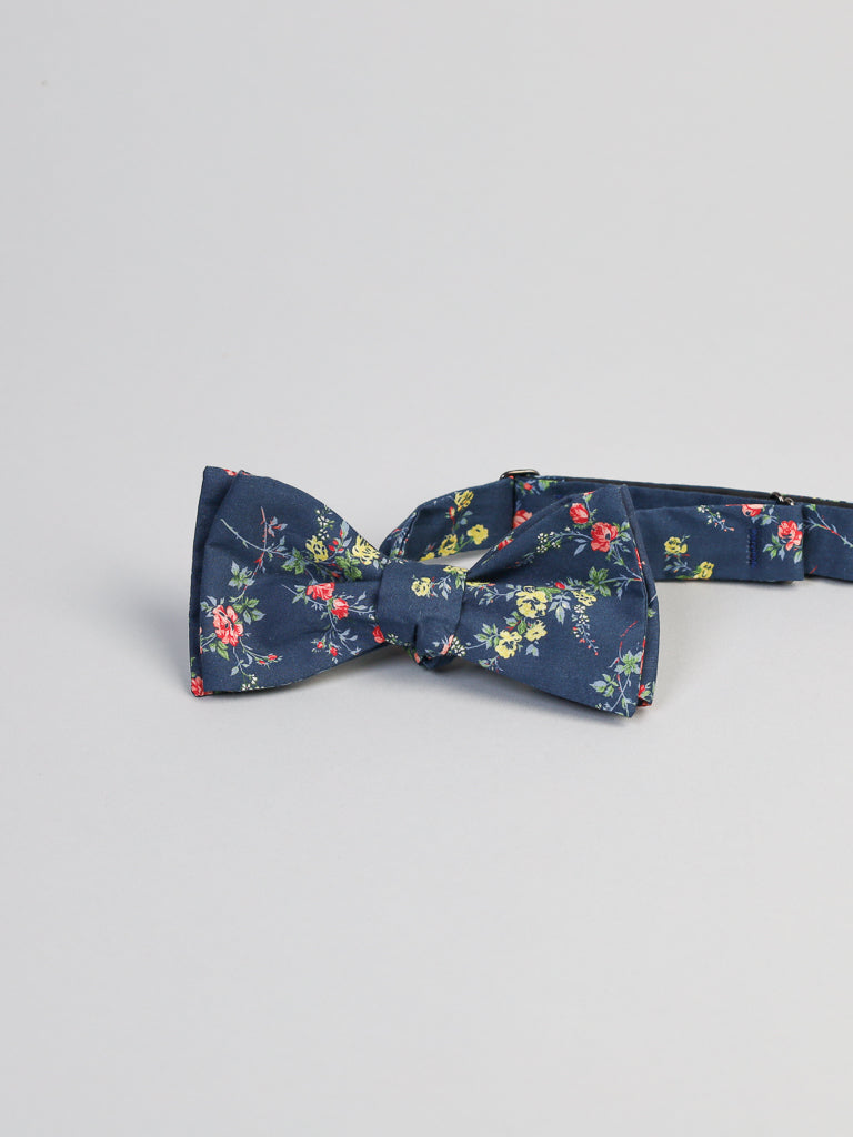 A navy blue bow tie featuring red, yellow, and green floral patterns on a light gray background, handcrafted in Brooklyn. Made from Liberty of Londons Tana Lawn cotton by ID Menswear, the Elizabeth version is elegantly designed with the band extending outwards.