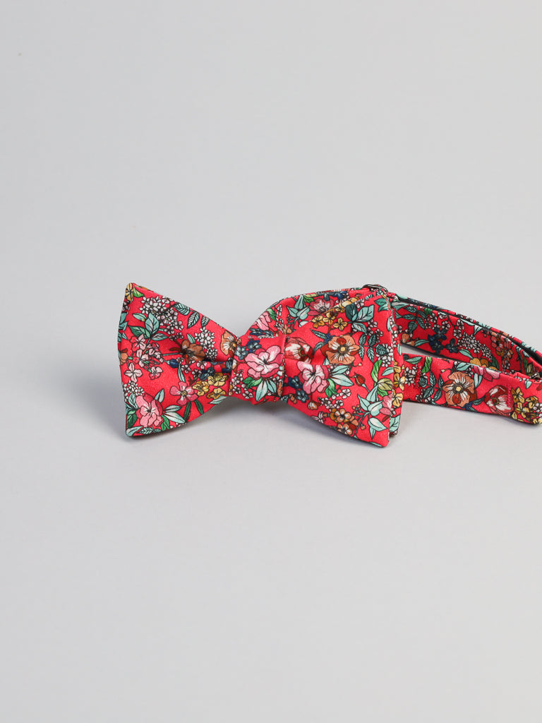 The Bouquet Cotton Bow Tie by ID Menswear features a red cotton base with green, white, and pink floral accents. Handmade in Brooklyn, it’s a unique semi-formal accessory, shown here on a plain light background.