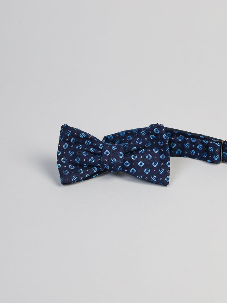 The Play Day Cotton Bow Tie by ID Menswear features a navy blue base with light blue floral patterns, handmade in Brooklyn and shown against a light gray background.