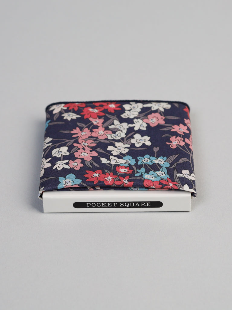 An ID Liberty of London Tana Lawn™ Sea Bloom Cotton Pocket Square by ID Menswear, showcasing white, red, and light blue flowers on a dark background, is neatly folded atop a white box labeled Pocket Square on a light gray surface.