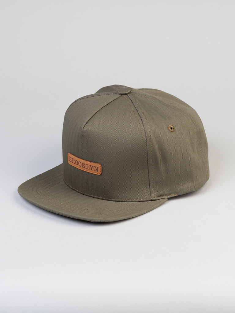 The Classic Cotton Herringbone Trucker by ID Menswear is an olive green baseball cap crafted from herringbone cotton, featuring a small brown patch with BROOKLYN on the front. It has a flat brim, a snapback closure, and is set against a plain gray background.