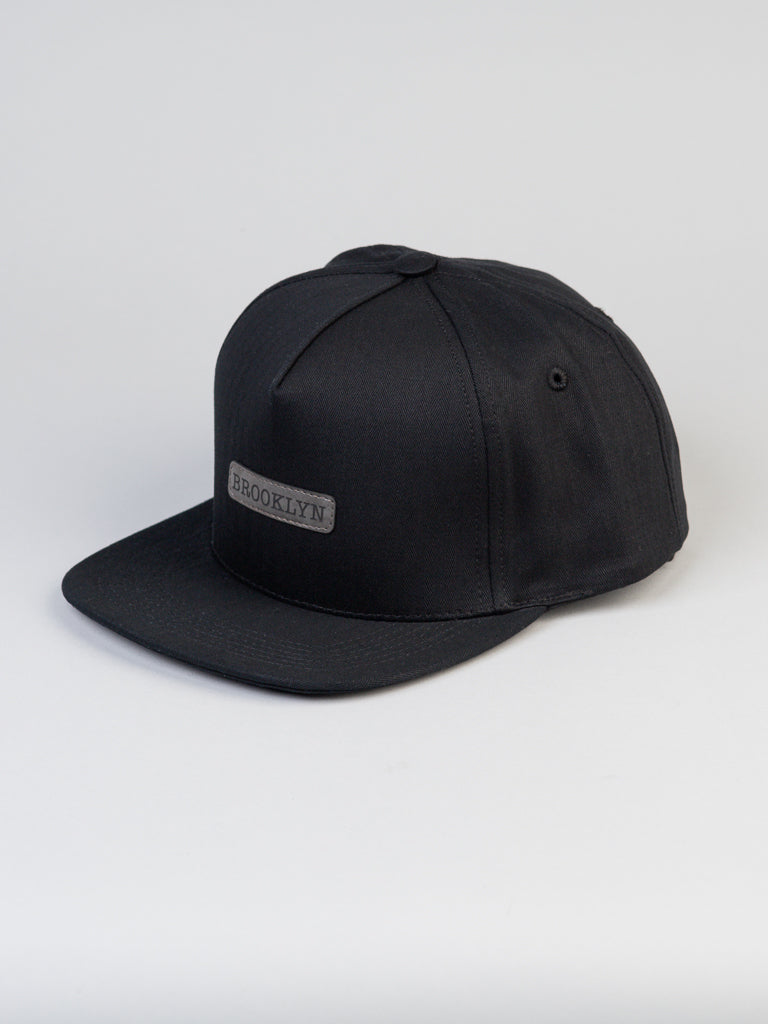The Classic Cotton Herringbone Trucker by ID Menswear is a stylish black cap with a flat brim, featuring a gray Brooklyn patch on the front panel. It has a snapback closure and is set against a plain white background for modern appeal.