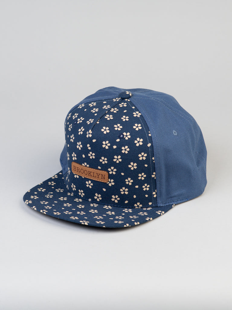 The ID Menswear Japanese Printed Sakura Cotton Trucker is a 100% cotton blue snapback cap featuring small white flowers on the front and flat brim, a brown leather BROOKLYN patch, and plain blue side panels.