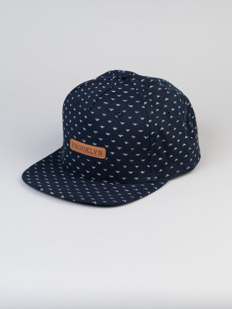 A dark blue Japanese Printed Nara Cotton Trucker features a small triangle pattern, a leather patch saying Brooklyn, and includes snapback closure with a curved brim. Its by ID Menswear and is set against a plain, light-colored background.