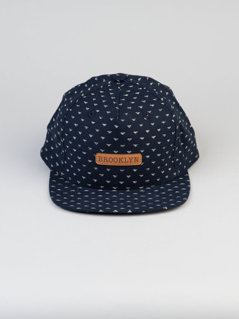 The Japanese Printed Nara Cotton Trucker by ID Menswear is a navy blue cap with a small triangular pattern and a brown leather BROOKLYN patch. It has a snapback closure on a clean white background.