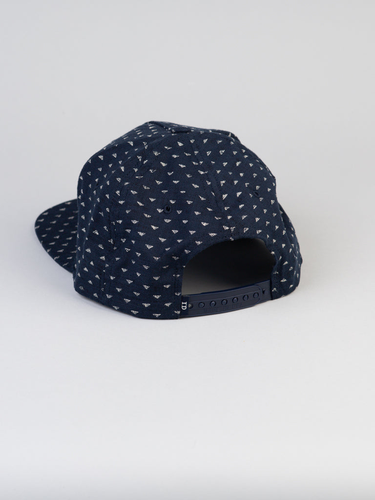 A Japanese Printed Nara Cotton Trucker by ID Menswear, dark blue with a small white triangle pattern, features an adjustable strap closure at the back. It is set against a plain white background.