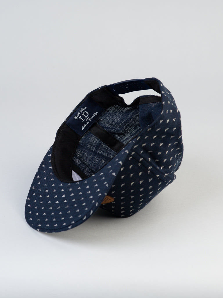 The Japanese Printed Nara Cotton Trucker by ID Menswear is a dark blue cap adorned with small white triangles and features a snapback closure. It embodies Brooklyns urban spirit, showcasing its checkered interior and black adjustable strap set to the side—an authentic ID Hat creation.