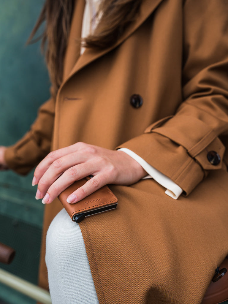 A person in a brown trench coat rests their hand on their knee, holding a Secrid Slimwallet Twist Twill Leather with RFID protection. Only the torso and hand are visible against a blurred background.