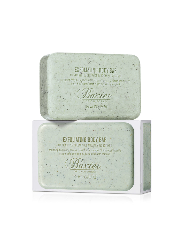 Two green Exfoliating Body Bars from Baxter are stacked, one resting sideways atop the other. The lower men's soap bar is partially wrapped in white packaging and scented with cedarwood and oakmoss.