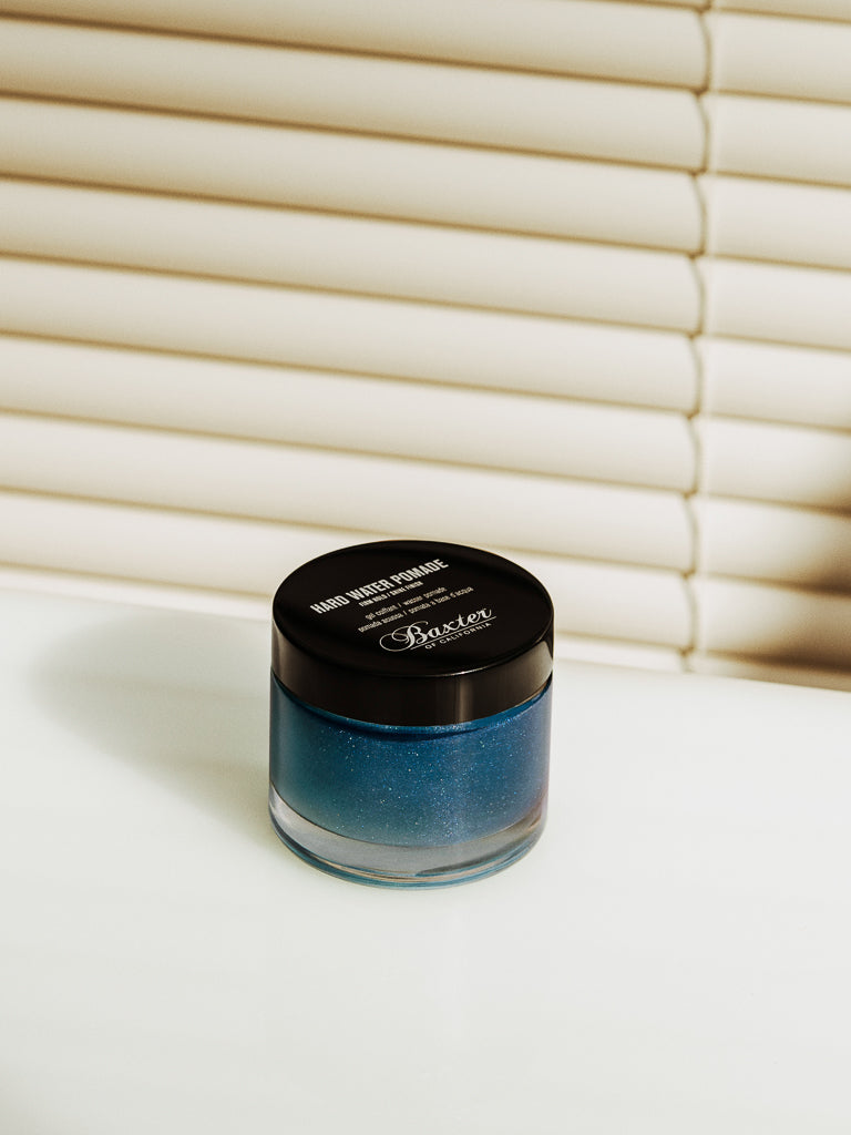 A small glass jar with a black lid labeled Baxter Hard Water Pomade Firm Hold and Shine Finish sits on a white surface before cream window blinds. The jar contains a shimmery blue, water-based formula for firm-hold, all-day hair control.