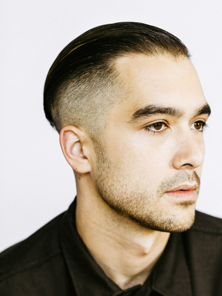A man with short, dark hair in a sleek side-parted undercut styled with Baxter’s Hard Water Pomade Firm Hold and Shine Finish, wearing a dark collared shirt, looks slightly to the side against a plain light background.