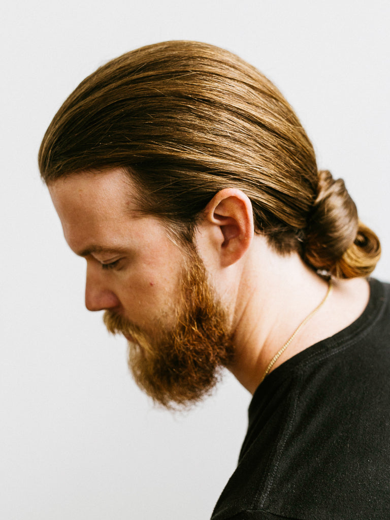 A man with light brown hair in a low bun and a full beard looks downward. His smooth, defined style suggests he used Baxter’s Cream Pomade Light Hold and Natural Finish. He wears a black shirt and thin gold necklace against a plain white background.