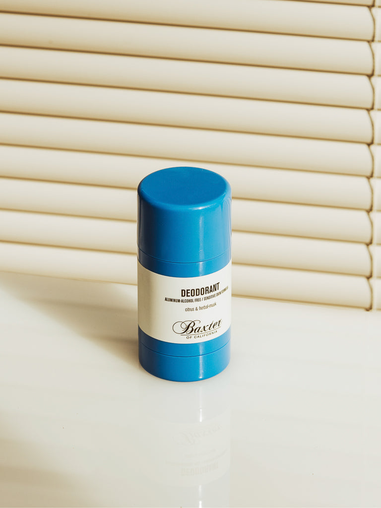 A bright blue Baxter Aluminium and Alcohol-Free Natural Deodorant stick is placed on a white surface in front of closed cream-colored blinds.