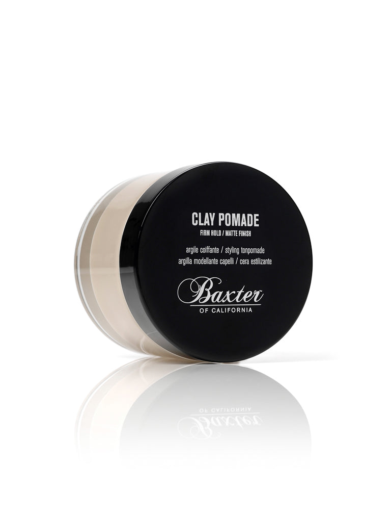 A round black and clear container of Baxter Clay Pomade Firm Hold and Matte Finish is shown on a white background, its white label text reflected slightly below.