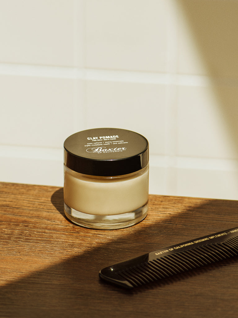 A glass jar of Baxter Clay Pomade Firm Hold and Matte Finish with a black lid sits on wood next to a black comb, against white tiles and bathed in warm sunlight.