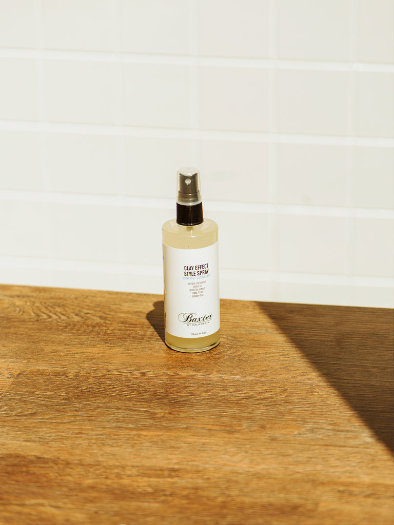 A bottle of Baxter’s Clay Effect Style Spray, valued for its matte finish, rests on a wooden surface bathed in sunlight before a backdrop of white tiled wall.
