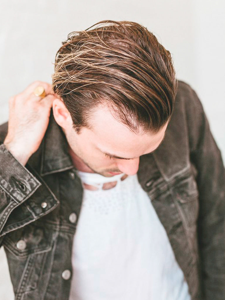 A man with slicked-back brown hair styled for a matte finish using Baxter’s Clay Effect Style Spray looks down, touching his neck. He wears a dark denim jacket over a white shirt against a light, blurred background.