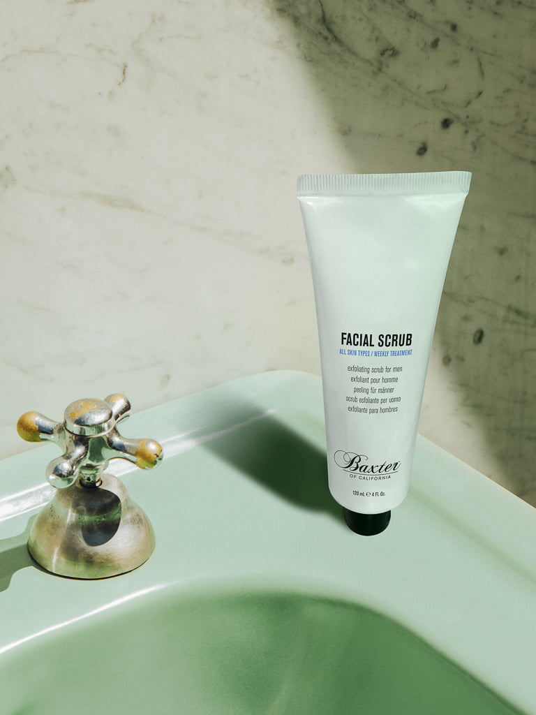 A tube of Baxter’s Exfoliating Facial Scrub to Tone and Smooth stands upright on a pale green sink by a silver faucet, sunlight highlighting its spot—ideal for exfoliating and helping prevent ingrown hairs.