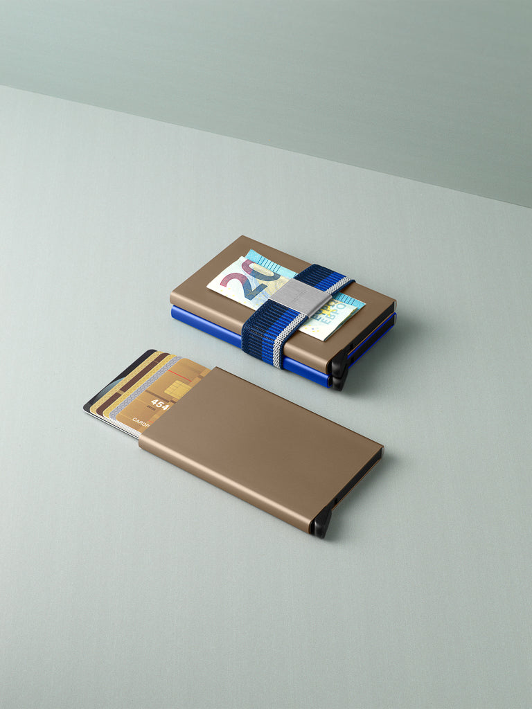 Two slim, metallic Secrid cardholders lie on a light gray surface. One uses the Money Band Cardprotector Add-on in blue to secure cash and cards; the other Cardprotector is partially open with several bank cards inside.